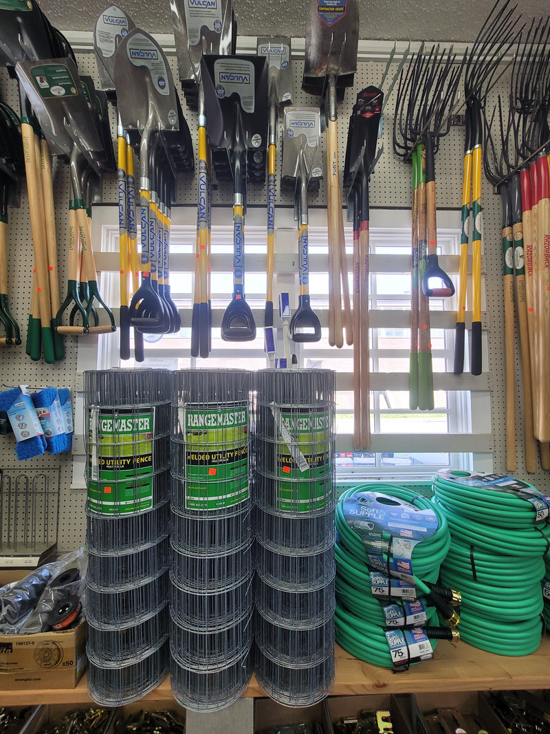 Wall displaying pitchforks, rakes, and a shelf with garden hoses and utility fence below