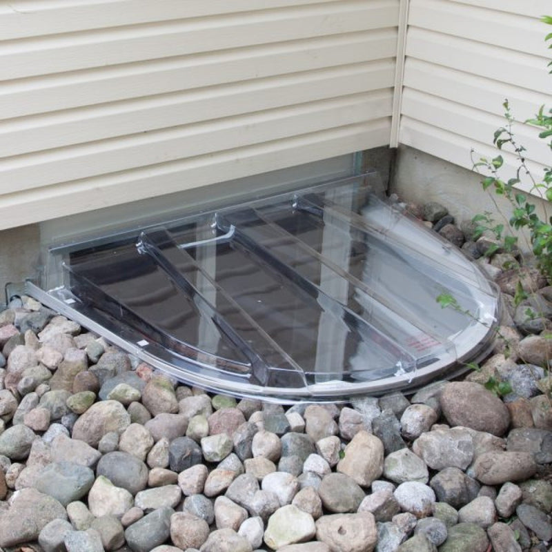 Clear plastic window well cover on rounded basement window, surrounded by beige siding and small rocks. Minimalist and functional outdoor scene.
